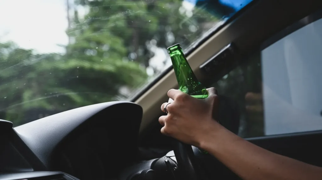 Man driving, holding green beer bottle inside car with trees outside