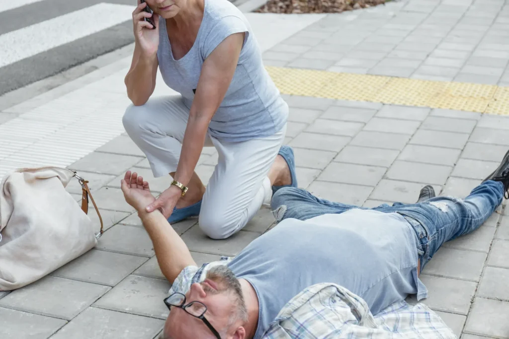 Senior kneels beside fainting person on sidewalk, providing outdoor first aid and support.