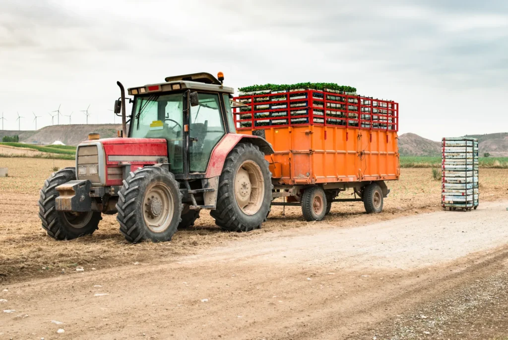 Orange off-road tractor hauling a loaded cage trailer on dusty rural dirt road